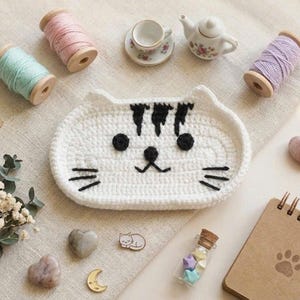 May include: A white crocheted cat-shaped tray with black yarn details for the eyes, nose, mouth, whiskers, and forehead stripes. The tray is surrounded by wooden spools of pastel yarn, a miniature tea set, heart-shaped stones, and a spiral notebook.