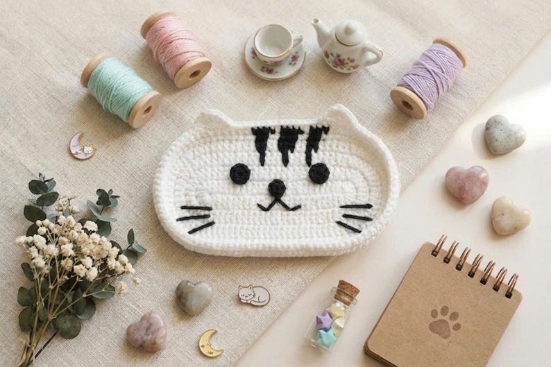 May include: A white crocheted cat-shaped tray with black yarn details for the eyes, nose, mouth, whiskers, and forehead stripes. The tray is surrounded by wooden spools of pastel yarn, a miniature tea set, heart-shaped stones, and a spiral notebook.