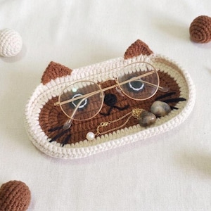 May include: An oval crochet tray shaped like a cat face with cream and brown yarn, featuring pointed ears, black embroidered whiskers, and a nose. A pair of gold-rimmed glasses, a delicate gold chain with a pearl, and two smooth stones rest inside.