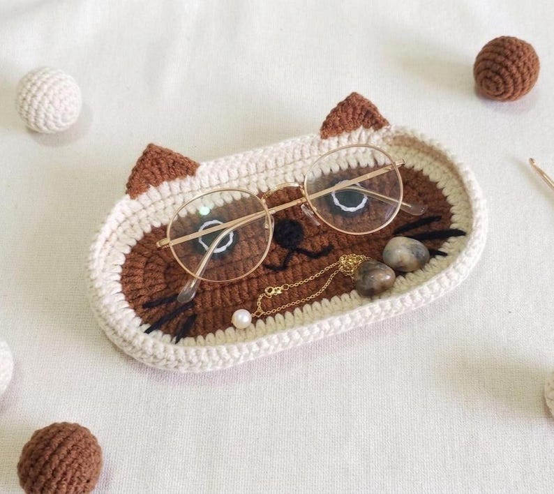 May include: An oval crochet tray shaped like a cat face with cream and brown yarn, featuring pointed ears, black embroidered whiskers, and a nose. A pair of gold-rimmed glasses, a delicate gold chain with a pearl, and two smooth stones rest inside.