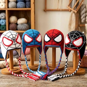 Puede incluir: Cuatro gorros de ganchillo con orejeras y borlas trenzadas, cada uno con un patr&oacute;n de telara&ntilde;a y grandes ojos blancos con bordes rojos. Los gorros son blancos, azules, rojos y negros, expuestos sobre soportes de madera con lazos.