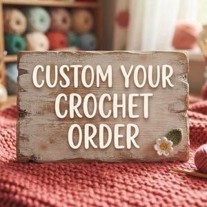 Pode incluir: Uma placa de madeira retangular com o texto CUSTOM YOUR CROCHET ORDER em letras brancas, decorada com uma pequena flor e uma folha de crochê. A placa repousa sobre um tecido rosa texturizado ao lado de um novelo de lã e uma agulha.