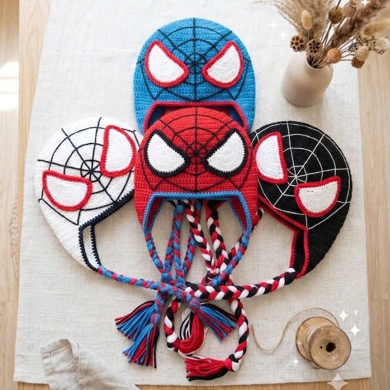 Puede incluir: Tres gorros de ganchillo con patr&oacute;n de telara&ntilde;a y grandes ojos blancos con bordes rojos. Los gorros son azules, rojos y negros, cada uno con largas orejeras trenzadas que terminan en borlas coloridas. El dise&ntilde;o imita una m&aacute;scara.