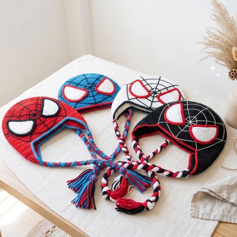 Puede incluir: Tres gorros de ganchillo con dise&ntilde;os inspirados en superh&eacute;roes. Uno es rojo con detalles azules, otro es azul con detalles rojos y otro es negro con detalles blancos. Cada gorro tiene parches oculares blancos, un patr&oacute;n de red y trenzas.