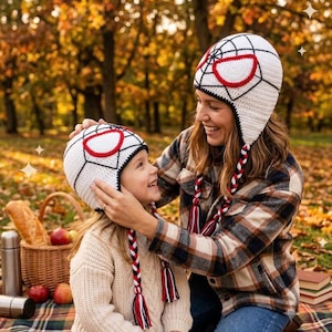 Puede incluir: Dos gorros de ganchillo blancos con patrones de telara&ntilde;a negros y parches oculares blancos con bordes rojos. Cada gorro incluye orejeras trenzadas largas en rojo, blanco y negro, terminadas con borlas a juego en los extremos.