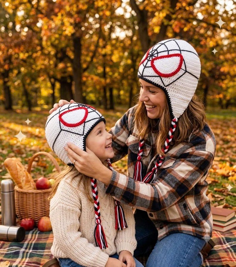 Puede incluir: Dos gorros de ganchillo blancos con patrones de telara&ntilde;a negros y parches oculares blancos con bordes rojos. Cada gorro incluye orejeras trenzadas largas en rojo, blanco y negro, terminadas con borlas a juego en los extremos.