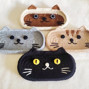 May include: Four oval-shaped crochet cat trays in different colours. One is brown and cream with dark ears, one is solid grey, one is tan with brown stripes, and one is solid black with bright yellow eyes. Each tray features embroidered whiskers.