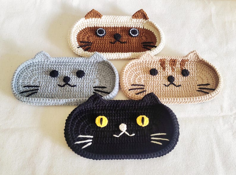 May include: Four oval-shaped crochet cat trays in different colours. One is brown and cream with dark ears, one is solid grey, one is tan with brown stripes, and one is solid black with bright yellow eyes. Each tray features embroidered whiskers.