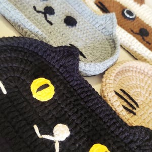 May include: A collection of four crocheted cat-shaped coasters or trays. The set includes a black cat with yellow eyes, a grey cat, a brown and white cat, and a tan tabby cat. Each piece features stitched facial details including eyes, noses, and whiskers.