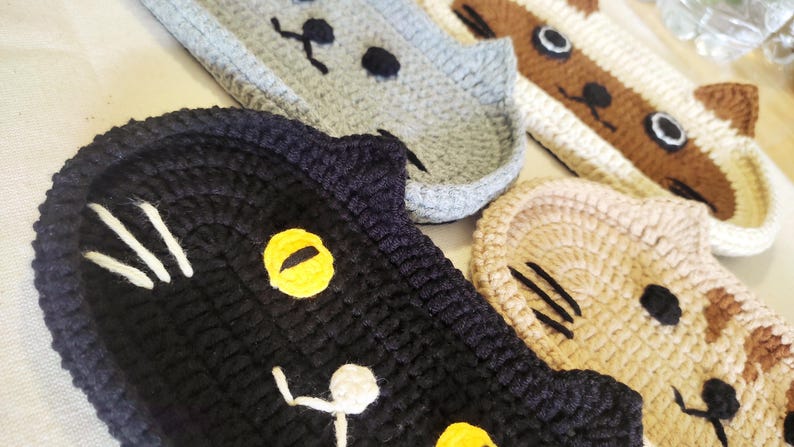 May include: A collection of four crocheted cat-shaped coasters or trays. The set includes a black cat with yellow eyes, a grey cat, a brown and white cat, and a tan tabby cat. Each piece features stitched facial details including eyes, noses, and whiskers.