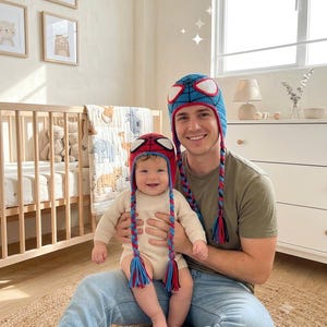 Puede incluir: Dos gorros de ganchillo a juego con un dise&ntilde;o en azul y rojo, detalles de telara&ntilde;a en negro y grandes parches blancos para los ojos. Cada gorro incluye largas orejeras trenzadas que terminan en borlas de colores, imitando una m&aacute;scara.