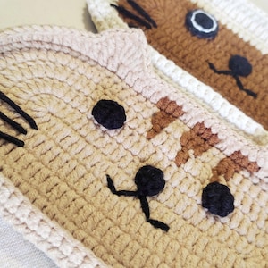 May include: Two handmade crochet cat-shaped coasters or mats. The front coaster is light beige with dark brown embroidered whiskers, black eyes, and a black nose and mouth. The rear coaster is brown and white with black eyes and whiskers.
