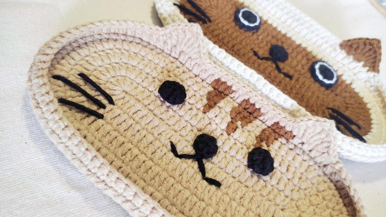 May include: Two handmade crochet cat-shaped coasters or mats. The front coaster is light beige with dark brown embroidered whiskers, black eyes, and a black nose and mouth. The rear coaster is brown and white with black eyes and whiskers.