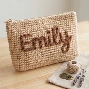 Peut inclure: Une pochette rectangulaire en crochet beige avec le prénom Emily écrit en lettres de laine marron en relief. La pochette est dotée d'une fermeture éclair dorée sur le bord supérieur. La texture est faite de mailles crochetées.