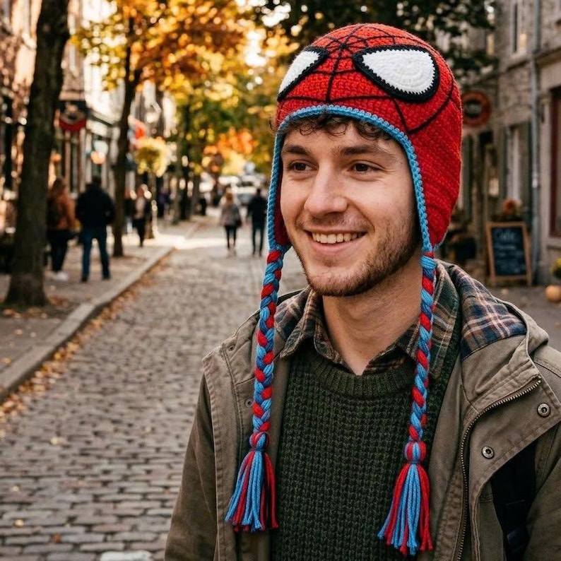 Puede incluir: Un gorro de ganchillo rojo y azul con un patr&oacute;n de telara&ntilde;a negro y grandes parches blancos en forma de ojos. El gorro cuenta con largas orejeras trenzadas de hilo rojo y azul que cuelgan a ambos lados y terminan en borlas a juego.