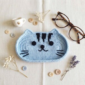 May include: A light blue crocheted cat-shaped tray with black yarn details for the eyes, nose, mouth, whiskers, and forehead stripes. The tray is surrounded by a small white ceramic cup, tortoiseshell glasses, several buttons, and dried flowers.