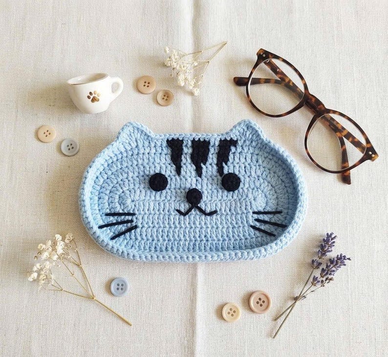 May include: A light blue crocheted cat-shaped tray with black yarn details for the eyes, nose, mouth, whiskers, and forehead stripes. The tray is surrounded by a small white ceramic cup, tortoiseshell glasses, several buttons, and dried flowers.