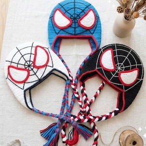 Puede incluir: Tres gorros de ganchillo con orejeras con dise&ntilde;os inspirados en superh&eacute;roes. Un gorro es azul, otro blanco y otro negro, cada uno con un patr&oacute;n de telara&ntilde;a, grandes ojos blancos con bordes rojos y largas trenzas con extremos de hilo.