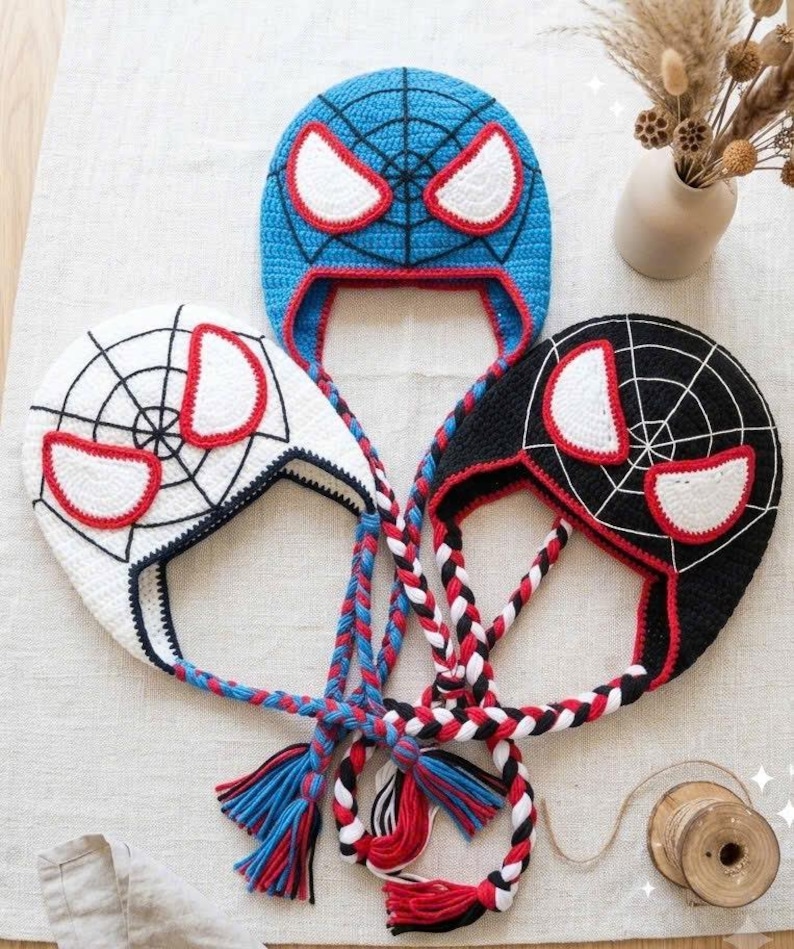 Puede incluir: Tres gorros de ganchillo con orejeras con dise&ntilde;os inspirados en superh&eacute;roes. Un gorro es azul, otro blanco y otro negro, cada uno con un patr&oacute;n de telara&ntilde;a, grandes ojos blancos con bordes rojos y largas trenzas con extremos de hilo.