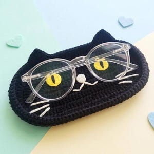 May include: A black crocheted cat-shaped tray featuring two yellow embroidered eyes, a small white nose, and white whiskers. A pair of clear-framed spectacles rests on top of the tray, positioned so the lenses align with the cat's eyes.