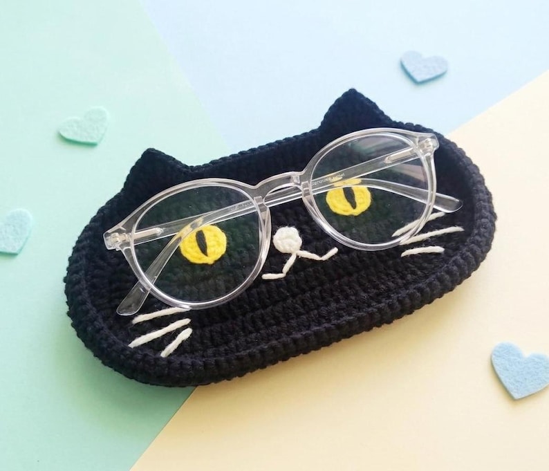 May include: A black crocheted cat-shaped tray featuring two yellow embroidered eyes, a small white nose, and white whiskers. A pair of clear-framed spectacles rests on top of the tray, positioned so the lenses align with the cat's eyes.