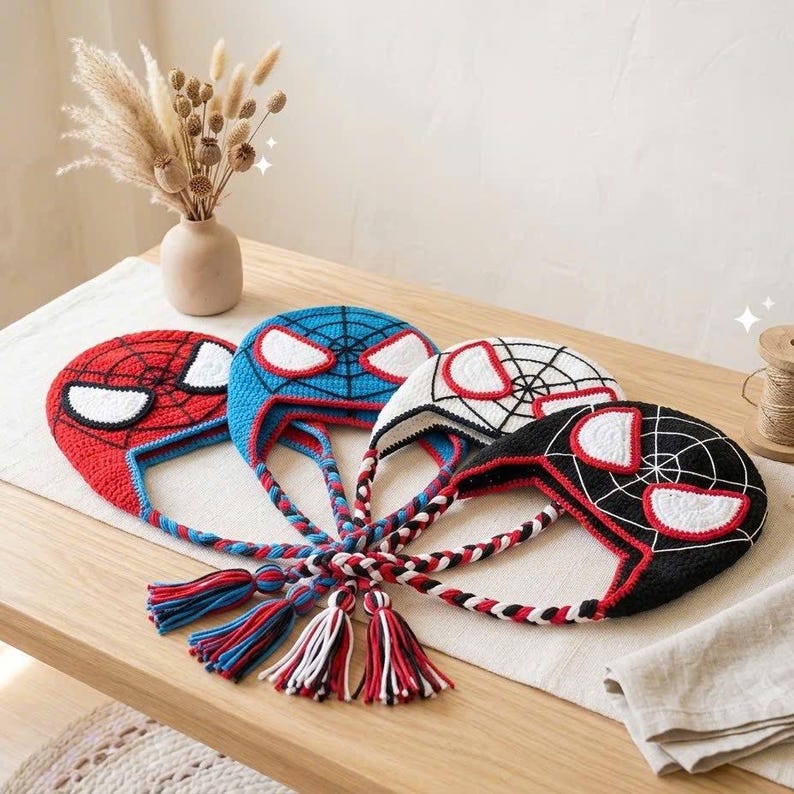 Puede incluir: Cuatro gorros de ganchillo con dise&ntilde;os inspirados en superh&eacute;roes, parches oculares blancos y patrones de telara&ntilde;a negros. Los gorros son rojos, azules, blancos y negros, con orejeras trenzadas que terminan en borlas de hilo multicolor.