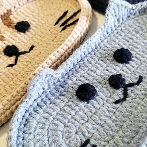 May include: A collection of four crocheted cat-shaped coasters in various colours including light grey, beige, brown, and black. Each coaster features embroidered black eyes, a nose, and whiskers, with the black cat having a yellow eye detail.