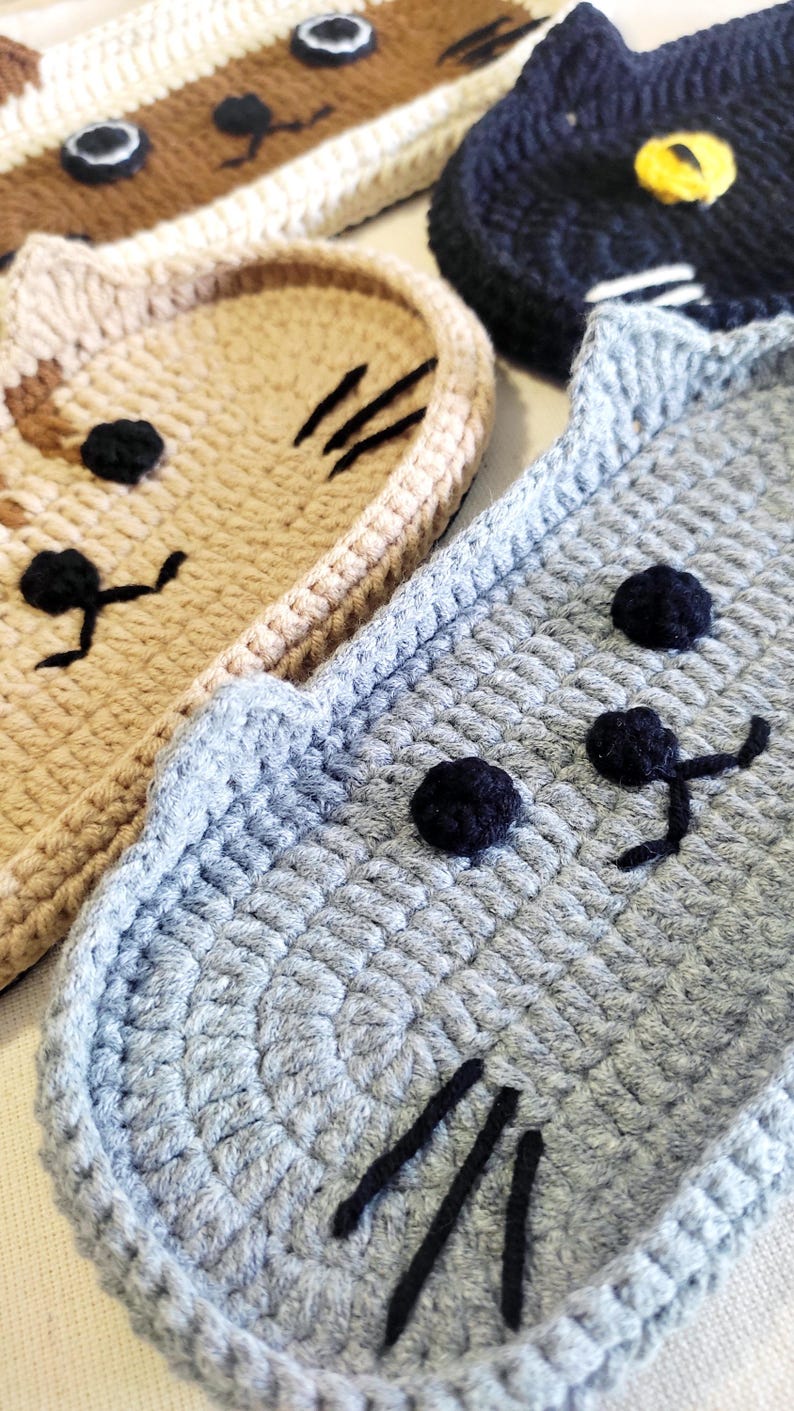 May include: A collection of four crocheted cat-shaped coasters in various colours including light grey, beige, brown, and black. Each coaster features embroidered black eyes, a nose, and whiskers, with the black cat having a yellow eye detail.