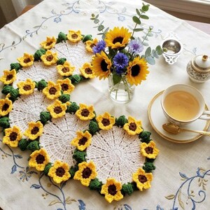 Könnte beinhalten: Gehäkelte Untersetzer mit gelben Sonnenblumenblüten und braunen Zentren. Eine Teetasse mit Tee, eine Teekanne und eine Vase mit Sonnenblumen und blauen Blumen sind auf einer Blumendecke zu sehen.