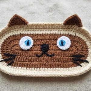 May include: An oval-shaped crochet tray featuring a brown cat face design with cream-coloured borders and ears. The cat has bright blue eyes with white circular accents, a small black nose, and black embroidered whiskers on both sides.