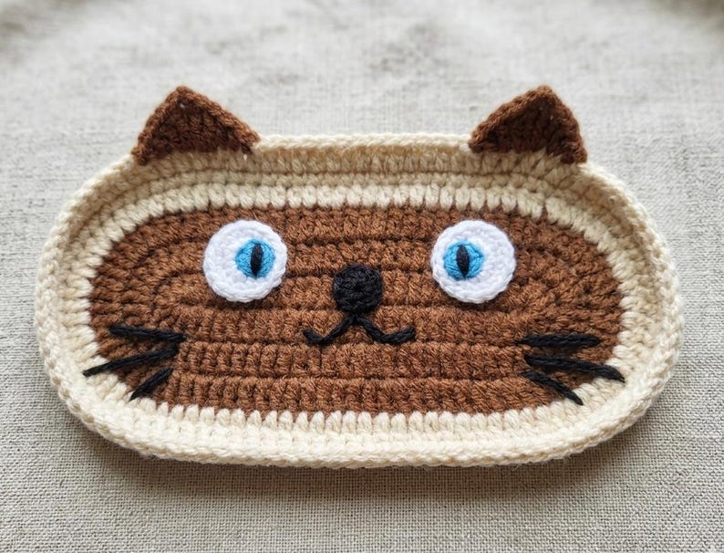 May include: An oval-shaped crochet tray featuring a brown cat face design with cream-coloured borders and ears. The cat has bright blue eyes with white circular accents, a small black nose, and black embroidered whiskers on both sides.
