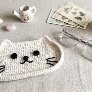 May include: A cream-coloured crochet tray shaped like a cat face, featuring two round black eyes, a small black nose, and three black embroidered whiskers on each side. The tray has a raised edge and is constructed from thick, textured yarn.