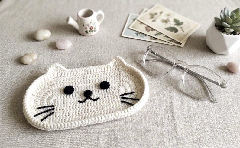 May include: A cream-coloured crochet tray shaped like a cat face, featuring two round black eyes, a small black nose, and three black embroidered whiskers on each side. The tray has a raised edge and is constructed from thick, textured yarn.