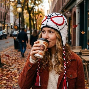 Puede incluir: Un gorro de ganchillo blanco con un patr&oacute;n de telara&ntilde;a negra y grandes parches oculares blancos con borde rojo. El gorro incluye dos largas trenzas laterales en rojo, blanco y negro, cada una terminada en una borla multicolor, sobre un su&eacute;ter beige.
