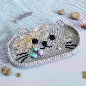 May include: A grey crocheted tray shaped like a cat face with black yarn whiskers, eyes, and a nose. The tray holds a pair of clear-framed glasses with a gold chain, a small turquoise fish charm, a purple bead, and a silver chain with a pearl.