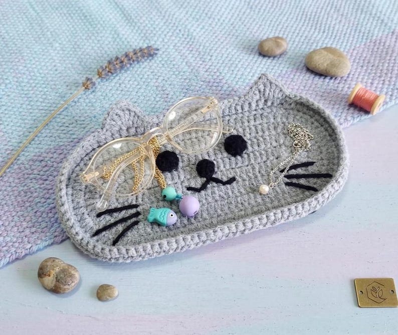 May include: A grey crocheted tray shaped like a cat face with black yarn whiskers, eyes, and a nose. The tray holds a pair of clear-framed glasses with a gold chain, a small turquoise fish charm, a purple bead, and a silver chain with a pearl.