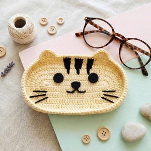 May include: A light yellow crochet tray shaped like a cat face with black yarn details for the eyes, nose, mouth, whiskers, and forehead stripes. The tray is oval-shaped with a raised edge, featuring a textured stitch pattern throughout the design.