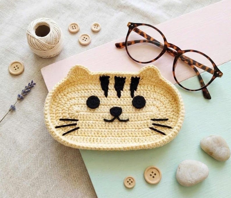 May include: A light yellow crochet tray shaped like a cat face with black yarn details for the eyes, nose, mouth, whiskers, and forehead stripes. The tray is oval-shaped with a raised edge, featuring a textured stitch pattern throughout the design.