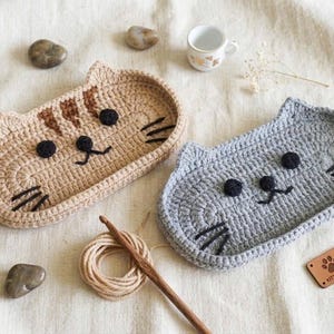 May include: Two oval crochet trays shaped like cat faces, one in tan with brown stripes and one in light grey. Both feature black yarn eyes, noses, and whiskers. A wooden crochet hook, a small ball of yarn, and a brown leather tag labelled KITTY CASE.