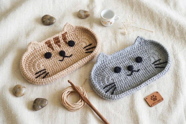 May include: Two oval crochet trays shaped like cat faces, one in tan with brown stripes and one in light grey. Both feature black yarn eyes, noses, and whiskers. A wooden crochet hook, a small ball of yarn, and a brown leather tag labelled KITTY CASE.