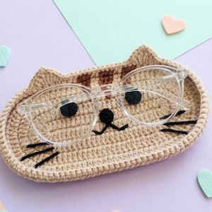 May include: A beige crocheted cat-shaped tray with brown striped ears, black yarn eyes, a black nose, and black whiskers. A pair of clear-framed spectacles rests on top of the tray, positioned so the lenses align with the cat's eyes.