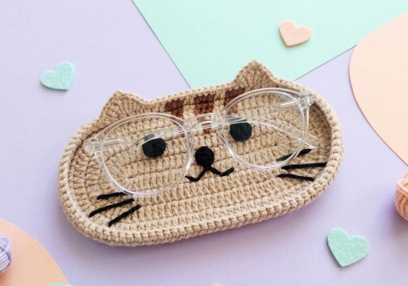 May include: A beige crocheted cat-shaped tray with brown striped ears, black yarn eyes, a black nose, and black whiskers. A pair of clear-framed spectacles rests on top of the tray, positioned so the lenses align with the cat's eyes.
