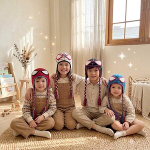 Puede incluir: Cuatro ni&ntilde;os con gorros de ganchillo con dise&ntilde;os de Spider-Man y orejeras trenzadas. Los gorros son rojos, blancos, negros y azules con grandes parches blancos para los ojos. Los ni&ntilde;os visten ropa de tonos neutros, incluyendo jers&eacute;is y petos.