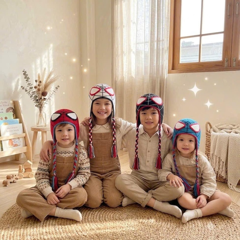 Puede incluir: Cuatro ni&ntilde;os con gorros de ganchillo con dise&ntilde;os de Spider-Man y orejeras trenzadas. Los gorros son rojos, blancos, negros y azules con grandes parches blancos para los ojos. Los ni&ntilde;os visten ropa de tonos neutros, incluyendo jers&eacute;is y petos.