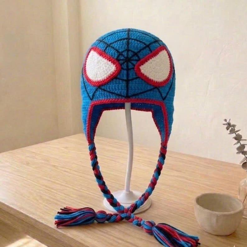 Puede incluir: Un gorro de ganchillo azul con un patr&oacute;n de telara&ntilde;a negra y grandes parches blancos para los ojos delineados en rojo. El gorro incluye dos orejeras trenzadas largas que cuelgan, cada una terminada en una borla multicolor de hilo azul, rojo y negro.