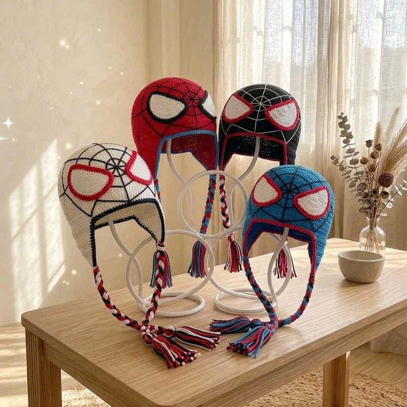 Puede incluir: Cuatro gorros de ganchillo con dise&ntilde;os inspirados en superh&eacute;roes, con grandes parches oculares blancos y patrones de telara&ntilde;a negros. Los gorros son rojos, negros, blancos y azules, con orejeras trenzadas largas y borlas de hilo.