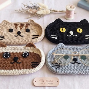 May include: Four oval-shaped crochet trays featuring cat faces in tan, black, brown, and grey. Each tray has embroidered eyes, noses, and whiskers. A pair of gold-rimmed glasses rests on the grey tray. A small leather tag reads "K&iacute;nh của bạn YOUR GLASSES."