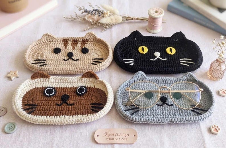 May include: Four oval-shaped crochet trays featuring cat faces in tan, black, brown, and grey. Each tray has embroidered eyes, noses, and whiskers. A pair of gold-rimmed glasses rests on the grey tray. A small leather tag reads "K&iacute;nh của bạn YOUR GLASSES."