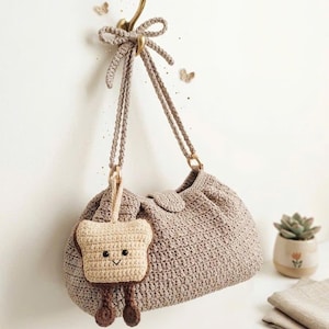 May include: A handmade crochet handbag in a neutral beige color, hanging from a gold hook. The bag features a matching strap and a small, smiling toast-shaped charm. The text "HANDMADE WITH LOVE" is visible at the bottom.