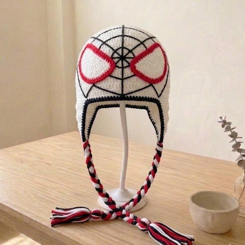 Puede incluir: Un gorro de ganchillo blanco con costuras negras en forma de telara&ntilde;a y dos grandes parches blancos para los ojos delineados en rojo. El gorro cuenta con orejeras y dos largas borlas trenzadas de hilo rojo, blanco y negro en los laterales.